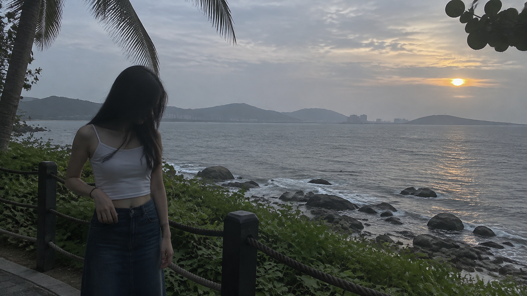 16:9 横屏，真实手机随手拍摄影，一位明确成年的年轻东亚女性站在海边观景步道旁，人物位于画面左侧约黄金分割位置，...