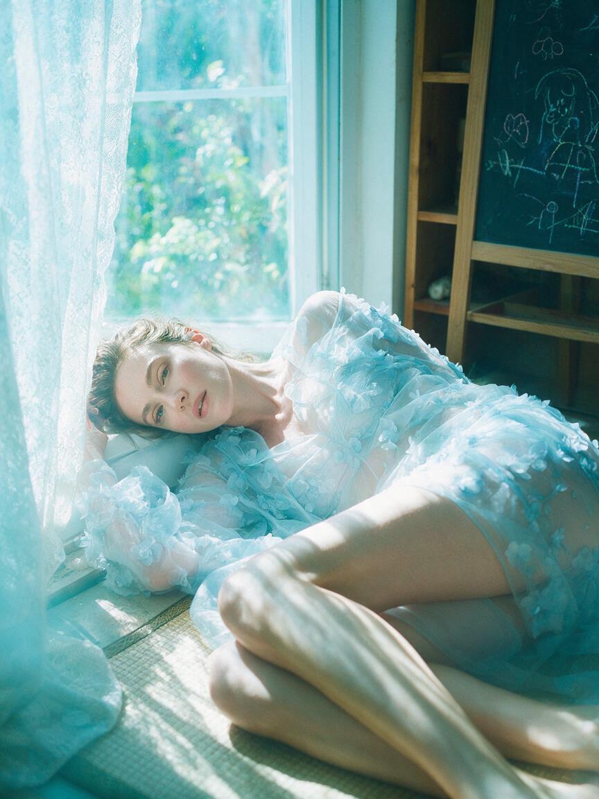 An adult woman reclines beside a bright window, gazing to...